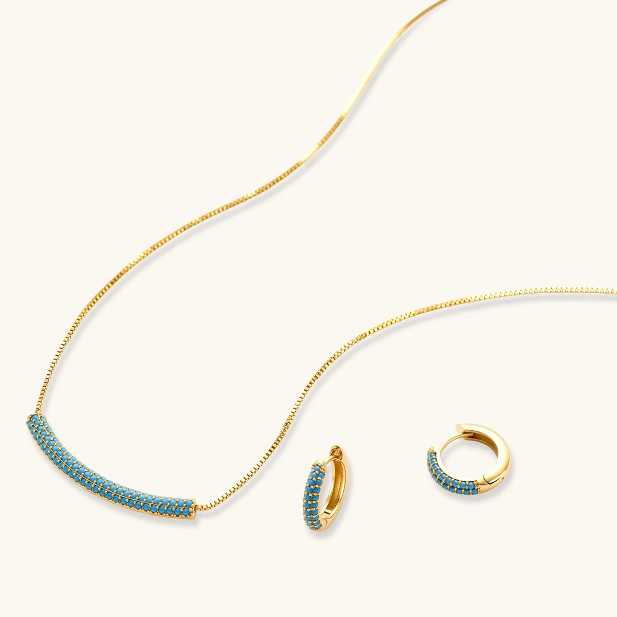Gold-plated curved necklace and hoop earrings with turquoise cubic zirconia stones on white background.